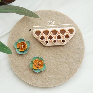 May include: A beige plastic flower petal cutter, measuring 5.18 cm, next to two small, colourful, handmade flowers. The flowers have teal petals and orange centres with gold accents. The cutter has multiple heart-shaped cutouts.