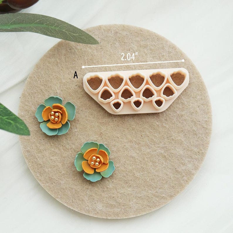 May include: A beige plastic flower petal cutter, measuring 5.18 cm, next to two small, colourful, handmade flowers. The flowers have teal petals and orange centres with gold accents. The cutter has multiple heart-shaped cutouts.