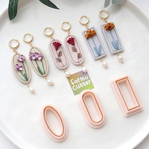 Puede incluir: Una colección de pendientes florales hechos a mano y cortadores de arcilla polimérica. Los pendientes presentan aros dorados, gotas de perlas y diseños florales coloridos. Los cortadores son ovalados y rectangulares, en color rosa claro. Incluye el texto "Formii Cutter".