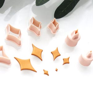May include: A collection of star-shaped cookie cutters and star-shaped cookies. The cutters are light pink, and the cookies are golden brown. The cutters are in various sizes and shapes, with some having a three-dimensional design.