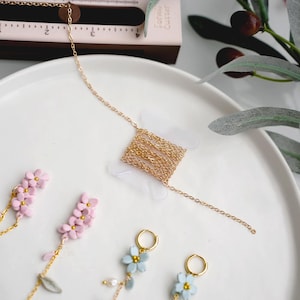 Puede incluir: Un collar dorado con un colgante cuadrado de cadenas entrelazadas. La imagen también muestra pendientes con colgantes de flores azules y rosas, todo sobre un plato blanco.