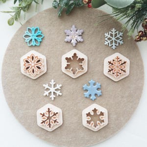 May include: A selection of snowflake-shaped cookie cutters and decorative snowflakes in various colours, including blue, purple, and white. The cutters are set on a neutral-toned surface, some within hexagonal frames. Festive and seasonal.