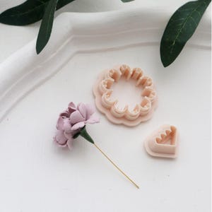 May include: A light pink flower and two flower-shaped cookie cutters. The flower has a green stem and a gold wire. The cookie cutters are in the shape of a flower and a heart with a scalloped edge. The items are on a white surface.