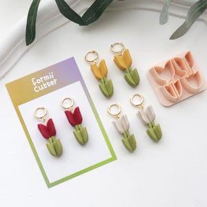 May include: Handmade tulip earrings in red, yellow, and white, with gold-coloured hoops. The earrings are displayed with a pink tulip-shaped cutter and a card that reads "formii Cutter". The earrings have green stems and leaves.