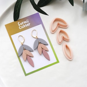 May include: A pair of leaf-shaped earrings in gray, peach, and mauve, with gold-colored ear wires, displayed with leaf-shaped clay cutters. The card reads "formii Cutter". The earrings and cutters are on a white surface.