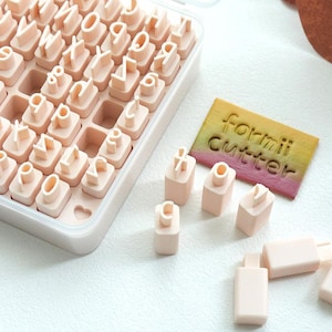 May include: A set of light pink alphabet letter stamps in a clear plastic case. The letters are arranged in rows and columns. A small wooden sign with the words "formii cutter" is also present.