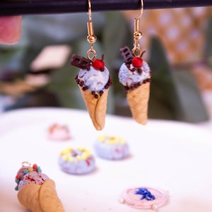 May include: Gold-toned dangle earrings featuring miniature ice cream cones. Each cone has a light blue scoop of ice cream, chocolate sprinkles, a chocolate bar, and a red cherry on top. Other miniature desserts are in the background.