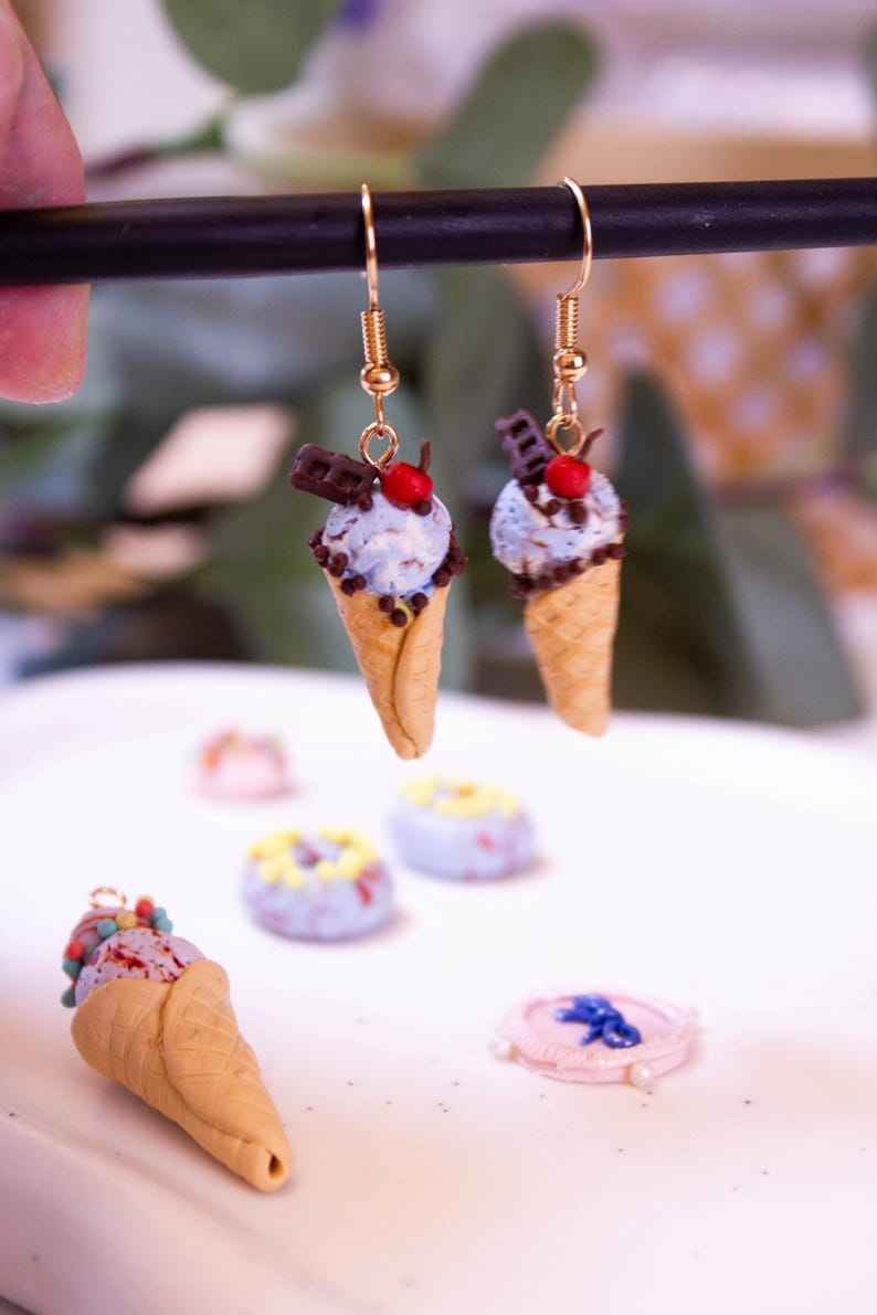 May include: Gold-toned dangle earrings featuring miniature ice cream cones. Each cone has a light blue scoop of ice cream, chocolate sprinkles, a chocolate bar, and a red cherry on top. Other miniature desserts are in the background.