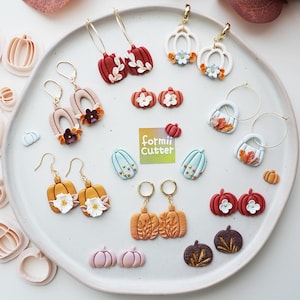 Könnte beinhalten: Eine Sammlung von Kürbis-Ohrringen aus Polymer-Ton und Ausstechformen auf einem Teller. Die Designs umfassen rote, weiße, orange, blaue und violette Kürbisse mit Blumenakzenten, Goldreifen und Steckern. Ein quadratisches Etikett in der Mitte zeigt formii cutter.