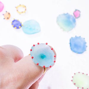 May include: A light blue ring with a gold band and a large, irregular-shaped stone. The stone is adorned with small red dots around the edge. Other similar, smaller, colorful stones are scattered in the background.