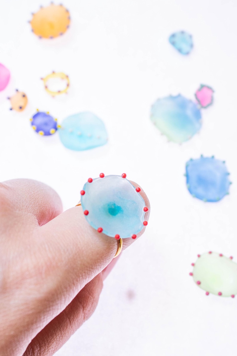 May include: A light blue ring with a gold band and a large, irregular-shaped stone. The stone is adorned with small red dots around the edge. Other similar, smaller, colorful stones are scattered in the background.