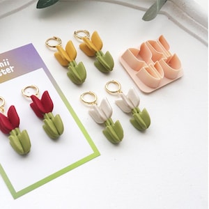 May include: Handmade tulip earrings in red, yellow, and white with gold-coloured hoops. The earrings have green stems and are displayed with a pink tulip-shaped cutter. The earrings are on a white surface.