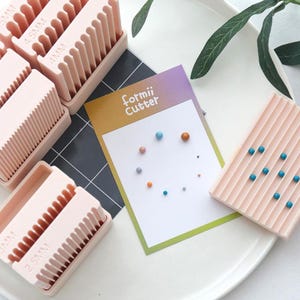 Puede incluir: Una colección de herramientas de manualidades de plástico rosa, incluyendo peines con diferentes tamaños de dientes, y una tarjeta con el texto "formii Cutter" y cuentas de colores. Las herramientas están dispuestas sobre una superficie blanca, sugiriendo un tema de manualidades.