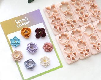 May include: A collection of colorful, fabric flower embellishments and flower-shaped cookie cutters. The embellishments are in various colors, including blue, yellow, and purple. The cookie cutters are arranged on pink rectangular molds. The text "formii Cutter" is visible.