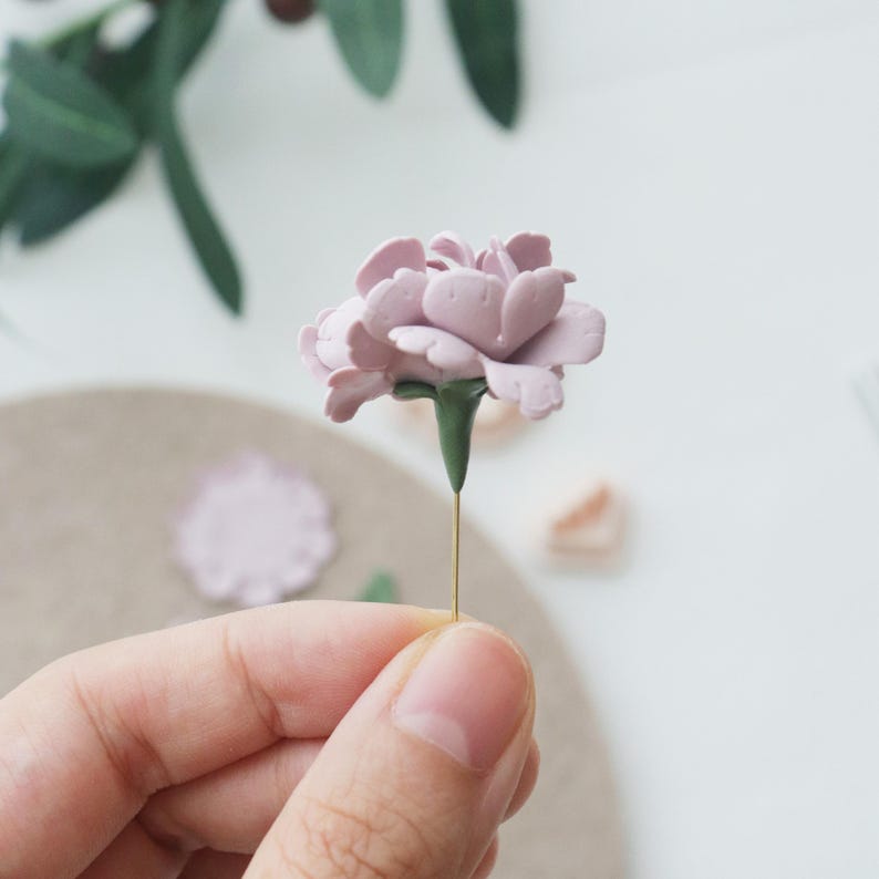May include: A delicate, handcrafted, light purple flower with green stem and a thin gold wire. The flower's petals are intricately detailed. The image is a close-up, showcasing the flower's texture and form. The flower is held by a person.