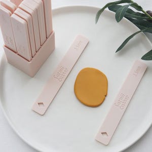 Puede incluir: Un conjunto de herramientas Formii Cutter de color rosa claro, que incluye varios cortadores rectangulares en un soporte y dos cortadores individuales. Los cortadores están etiquetados con "3,5 mm" y el nombre de la marca. También hay un pequeño trozo de arcilla amarilla.