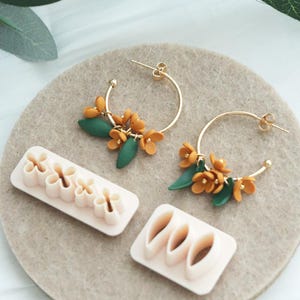 Op de afbeelding: Gouden oorringen met oranje bloem- en groene bladaccenten. Twee sets oorbellen worden getoond met twee sets roze bloemvormige kleisnijders. De oorbellen en snijders liggen op een neutrale ondergrond.