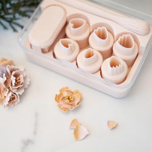 May include: A light pink flower-making kit with various heart-shaped cutters, a small rolling pin, and a rectangular tool. The kit is in a clear plastic container. A finished flower and loose petals are also visible.