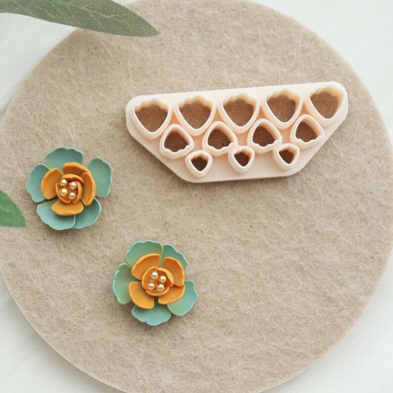 May include: A light beige craft mat with a flower-shaped cutter and two decorative flowers. The cutter is light pink and has multiple flower-shaped cutouts. The flowers are teal, orange, and gold, with small gold bead centres.