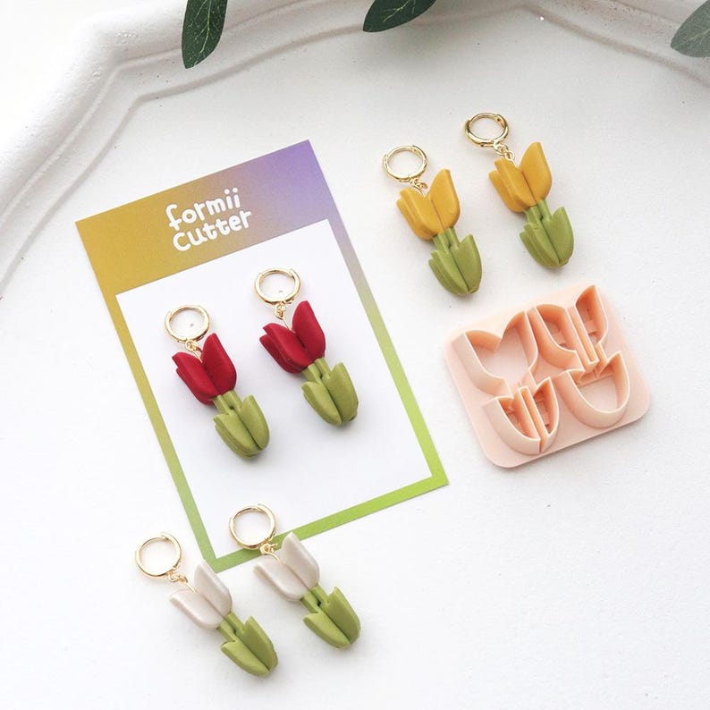 May include: Handmade tulip earrings in red, yellow, and white with green stems and gold hoops. A pink Formii Cutter is also visible. The earrings are displayed on a white surface with a decorative border.