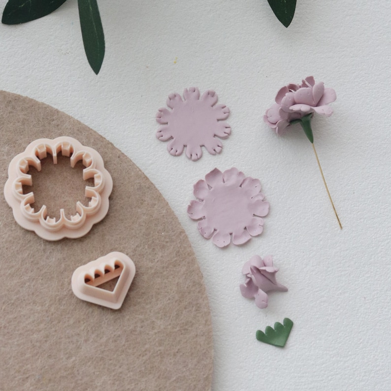 May include: A collection of light pink flower-shaped cookie cutters and flower petals. The cutters are in various shapes, including a scalloped circle and a triangle. A finished flower with a green stem is also present, along with a few green leaf cutouts.