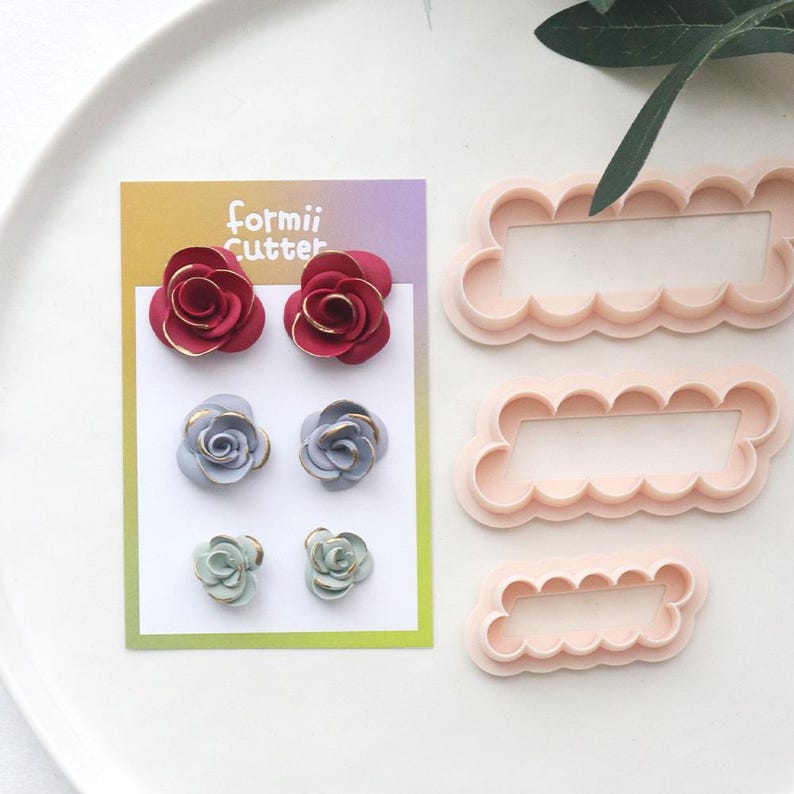 May include: A selection of craft supplies featuring floral designs. The image shows three pairs of rose-shaped embellishments in red, blue, and green, with gold detailing. There are also three pink, scalloped-edge frame cutters. The card reads "formii Cutter".
