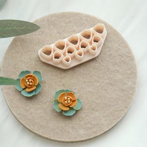 May include: A light beige flower-shaped cookie cutter set with various sizes, and two handmade flowers. The flowers have teal petals and orange centres with small gold beads. The items are arranged on a beige felt circle.