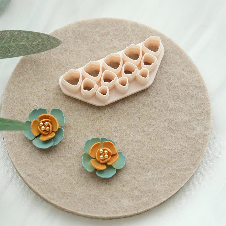 May include: A light beige flower-shaped cookie cutter set with various sizes, and two handmade flowers. The flowers have teal petals and orange centres with small gold beads. The items are arranged on a beige felt circle.