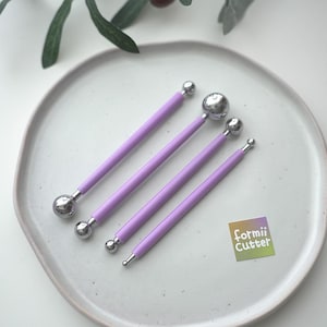 May include: A set of four double-ended ball stylus tools with lavender-colored handles and polished silver metal spheres of varying sizes. A small square sticker with the text formii Cutter is positioned near the tools on the surface.