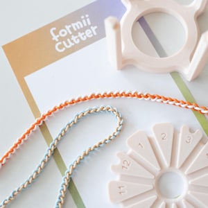May include: A craft image featuring a "formii Cutter" tool, with two braided cords in orange/white and blue/beige. A pink circular tool with numbered slots and a pink frame are also visible. The background is white and gradient.