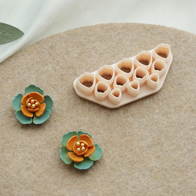 May include: Two handmade, colourful flower embellishments with teal petals and orange centres with gold beads. A light peach-coloured plastic flower-shaped cutter with multiple petal shapes is also shown. The items are on a neutral-toned surface.