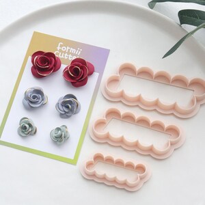 May include: An assortment of craft supplies, including floral decorations and cookie cutters. The image shows small, decorative flowers in red, blue, and green, with gold detailing. Next to them are three pink, scalloped-edge rectangular cutters. The card reads "formii Cutte".