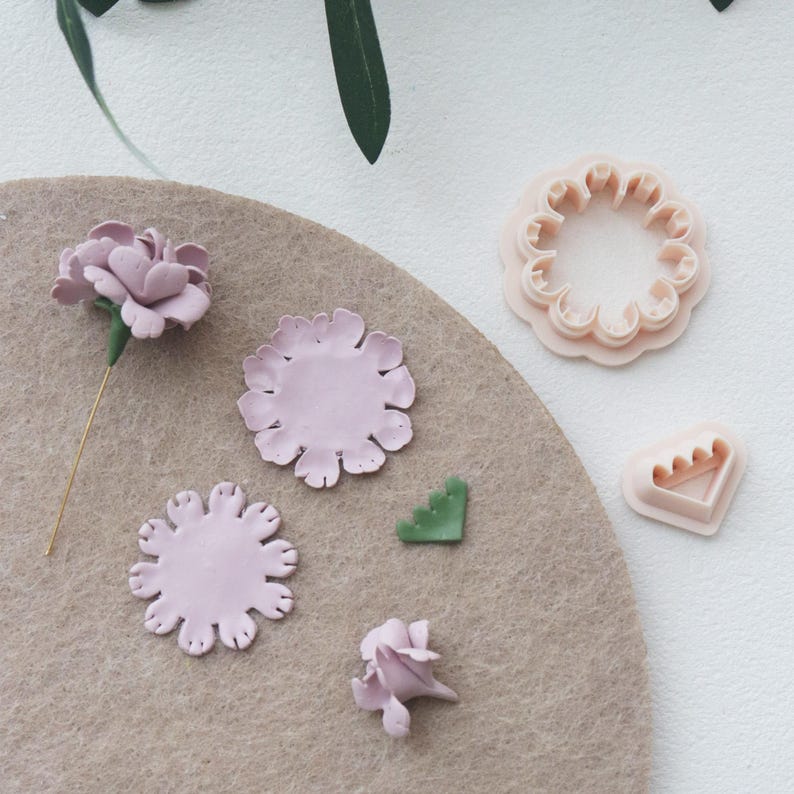 May include: A collection of light pink flower-shaped clay pieces and tools. The arrangement includes a completed flower, several petal cutouts, a green leaf, and two flower-shaped cutters. The items are arranged on a tan felt surface.