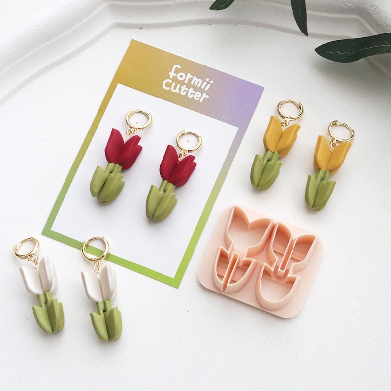 May include: Handmade tulip earrings in red, yellow, and white, with gold hoops. The earrings are displayed with a pink tulip-shaped cookie cutter and a card that reads "formii Cutter".
