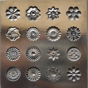 May include: Sixteen silver-coloured metal flower-shaped embellishments. Each flower has a unique design, with varying petal shapes and centre details. The embellishments are arranged on a textured, silver-toned metal surface.