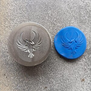 May include: A round, silver-colored metal object with a detailed bird design, next to a small, blue, rubbery object with the same bird design. The objects are on a gray surface.