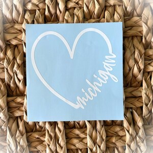 May include: A light blue sticker featuring a white heart outline with the word "Michigan" in a cursive font. The sticker is set against a woven, light brown background.
