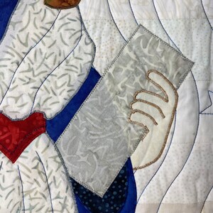 May include: A close-up of a quilt with a white background and blue, red, and white fabric pieces. The quilt features a hand holding a piece of fabric with a star design.