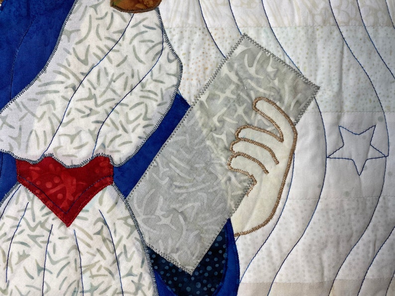 May include: A close-up of a quilt with a white background and blue, red, and white fabric pieces. The quilt features a hand holding a piece of fabric with a star design.
