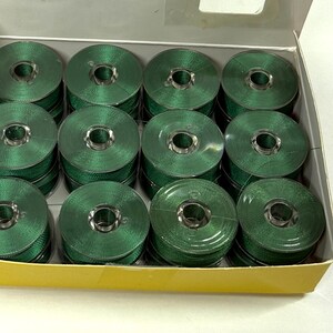 May include: A white cardboard box containing twelve circular sewing machine bobbins, each wound with dark green thread. The bobbins are arranged in three rows of four, featuring a metallic center core and a smooth, uniform green thread finish.