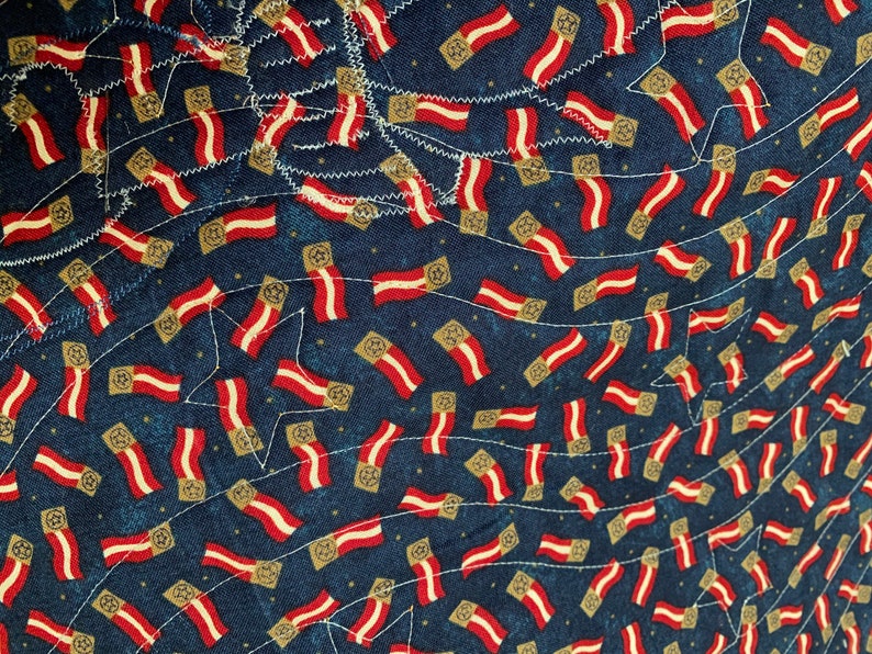 May include: A blue fabric with a repeating pattern of red, white, and gold flags. The flags are arranged in a diagonal pattern and are sewn onto the fabric.