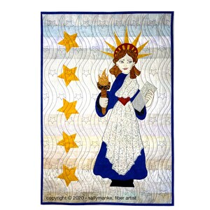 May include: A quilted wall hanging featuring a depiction of the Statue of Liberty, holding a torch and a document. The background is a blue and white pattern with five yellow stars.