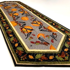 May include: A rectangular table runner with a gray center featuring orange pumpkins, black crows, and fall foliage. The border is black with green and brown leaves. The runner is quilted.