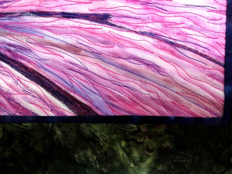May include: A close-up of a pink and purple fabric with a dark blue border. The fabric has a quilted design with pink and purple stitching.