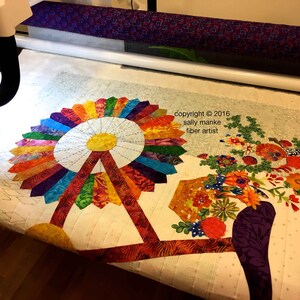 Puede incluir: Una colcha colorida con un dise&ntilde;o de molino de viento y una cesta de flores. La colcha se est&aacute; acolchando en una m&aacute;quina de acolchado de brazo largo. El texto "copyright &copy; 2016 sally manke fiber artist" es visible en la esquina superior derecha de la imagen.