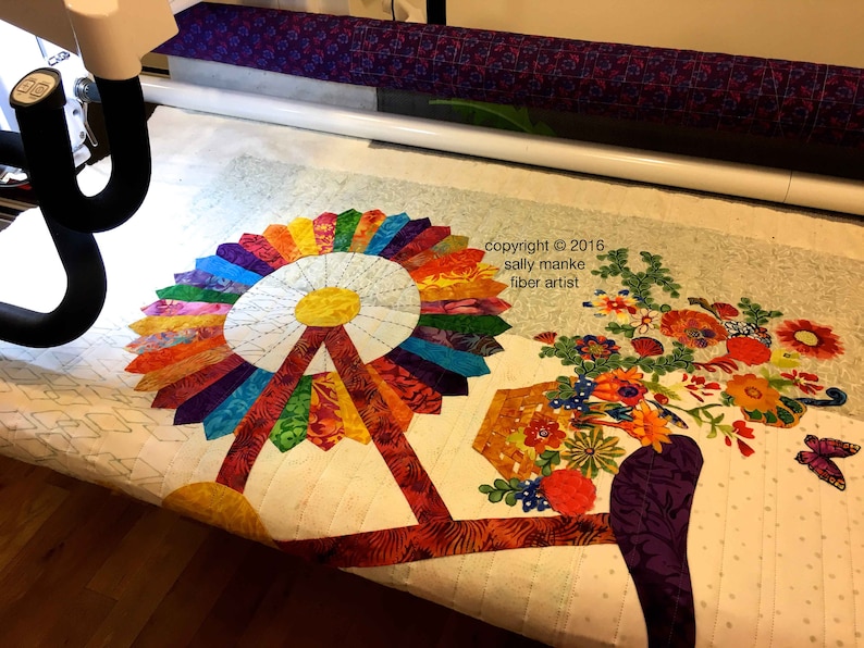 Puede incluir: Una colcha colorida con un dise&ntilde;o de molino de viento y una cesta de flores. La colcha se est&aacute; acolchando en una m&aacute;quina de acolchado de brazo largo. El texto "copyright &copy; 2016 sally manke fiber artist" es visible en la esquina superior derecha de la imagen.