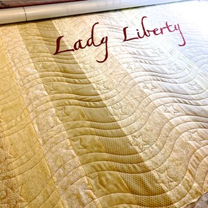 May include: A beige and cream quilt with a wavy pattern and stars. The quilt is labeled "Lady Liberty" in red lettering.