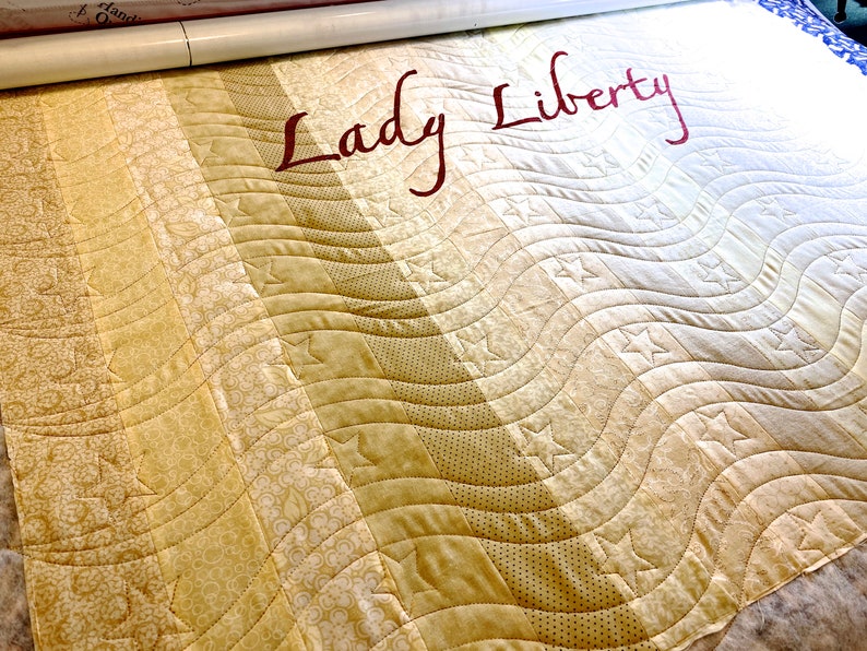 May include: A beige and cream quilt with a wavy pattern and stars. The quilt is labeled "Lady Liberty" in red lettering.