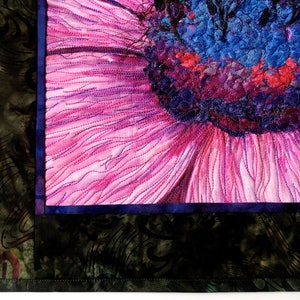 May include: Close-up of a pink flower with a blue and purple centre. The flower is quilted with intricate stitching. The background is a dark green fabric with a floral pattern.