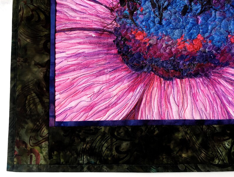 May include: Close-up of a pink flower with a blue and purple centre. The flower is quilted with intricate stitching. The background is a dark green fabric with a floral pattern.